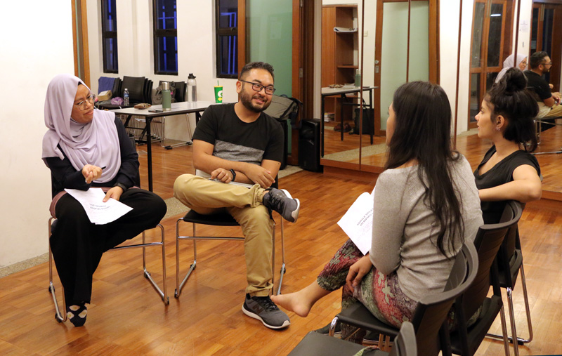 Four persons seated on chairs in the 42 Waterloo Street Meeting Room, talking to one another. Two hold scripts in their hands.