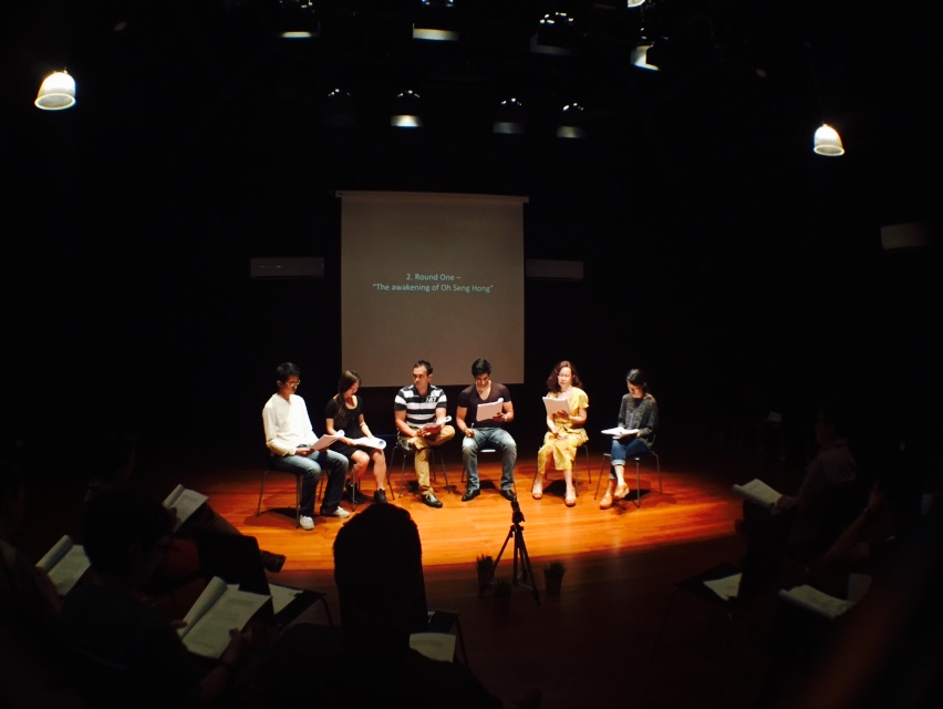 Six persons seated in a row in a black box, reading from scripts. Behind them is a screen with the words '2. Round One - 'The awakening of Oh Seng Hong' projected on it.