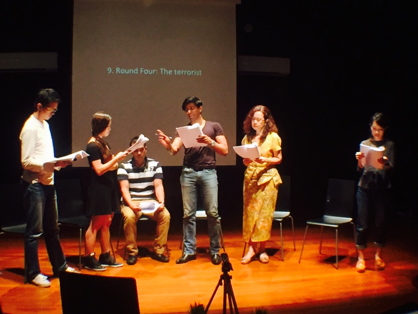 Six persons in a black box, five standing in front of their chairs, one seated, speaking while reading their scripts. Behind them is a screen with the words '9. Round Four - The terrorist' projected on it.