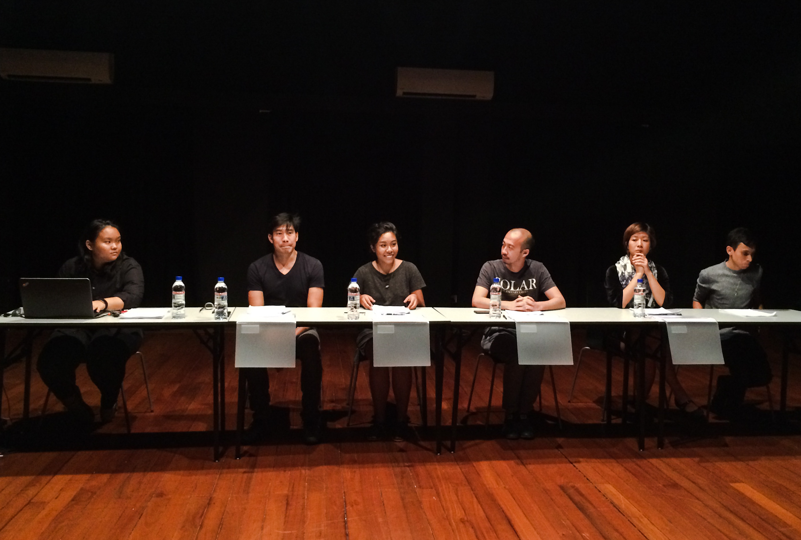 Six persons seated in a row at a table, with scripts in front of them. Four plastic folders hang over the edge of the table in front of them.