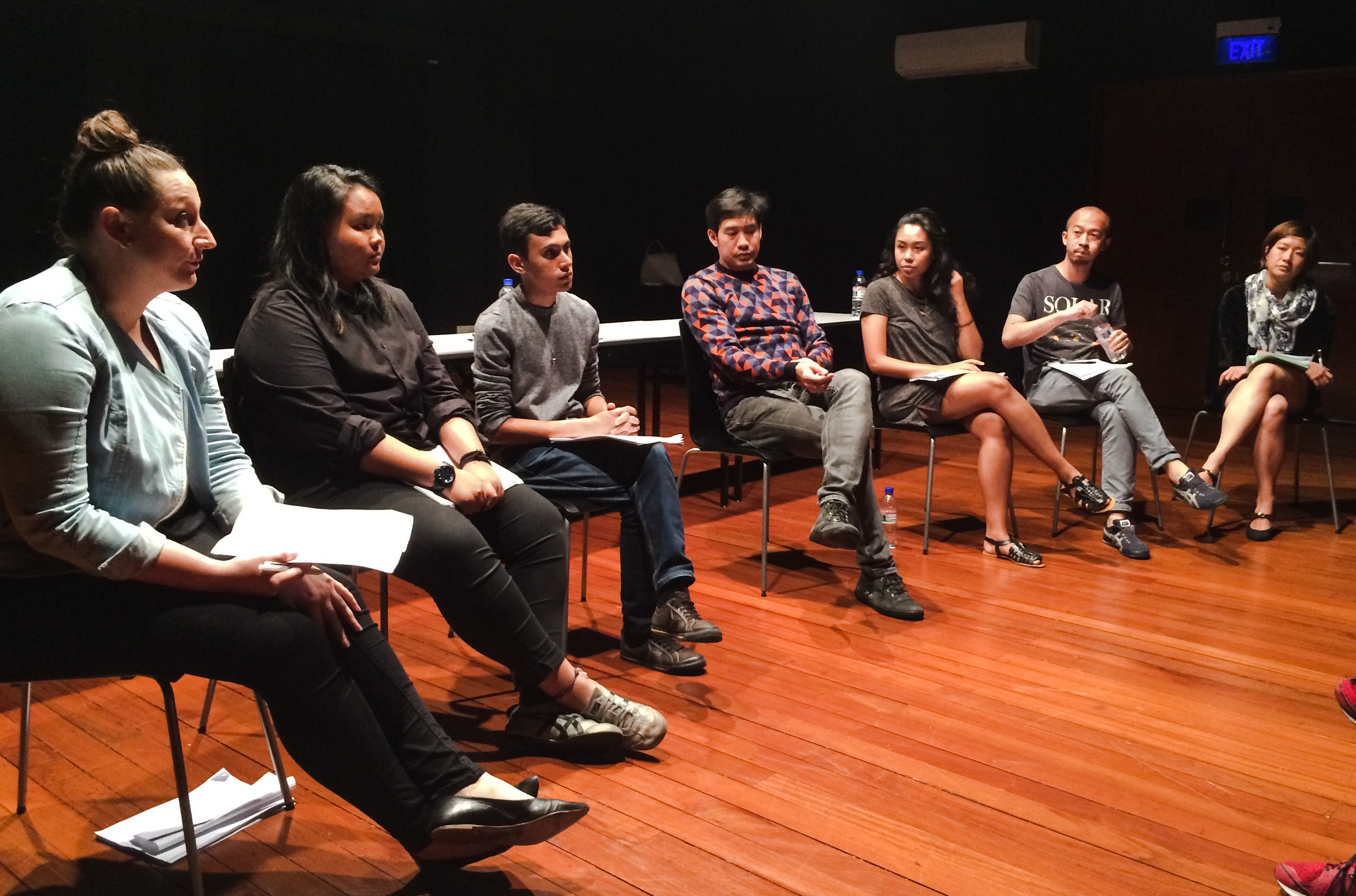 Seven people seated in a row of chairs in a black box.