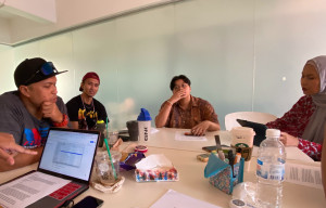 A group of four persons seated around a table covered with pencil cases, water bottles and a laptop.