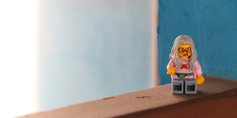 A Lego figure with long grey hair, wearing red glasses and a pink sweater, on a brown railing.