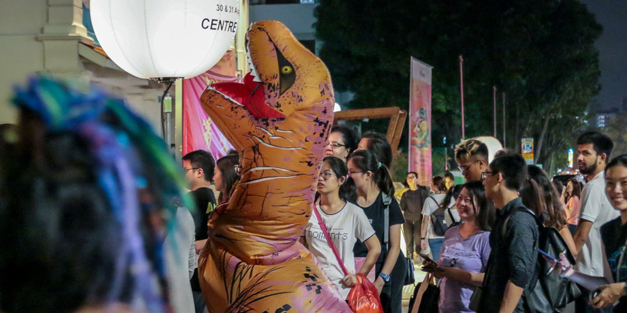 A crowd of people outside 42 Waterloo Street, at night. Amidst them, front and centre, is a person in a dinosaur costume.