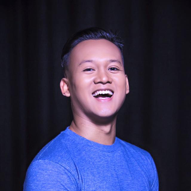 Coloured headshot of Kaykay Nizam smiling widely, dressed in a blue shirt.