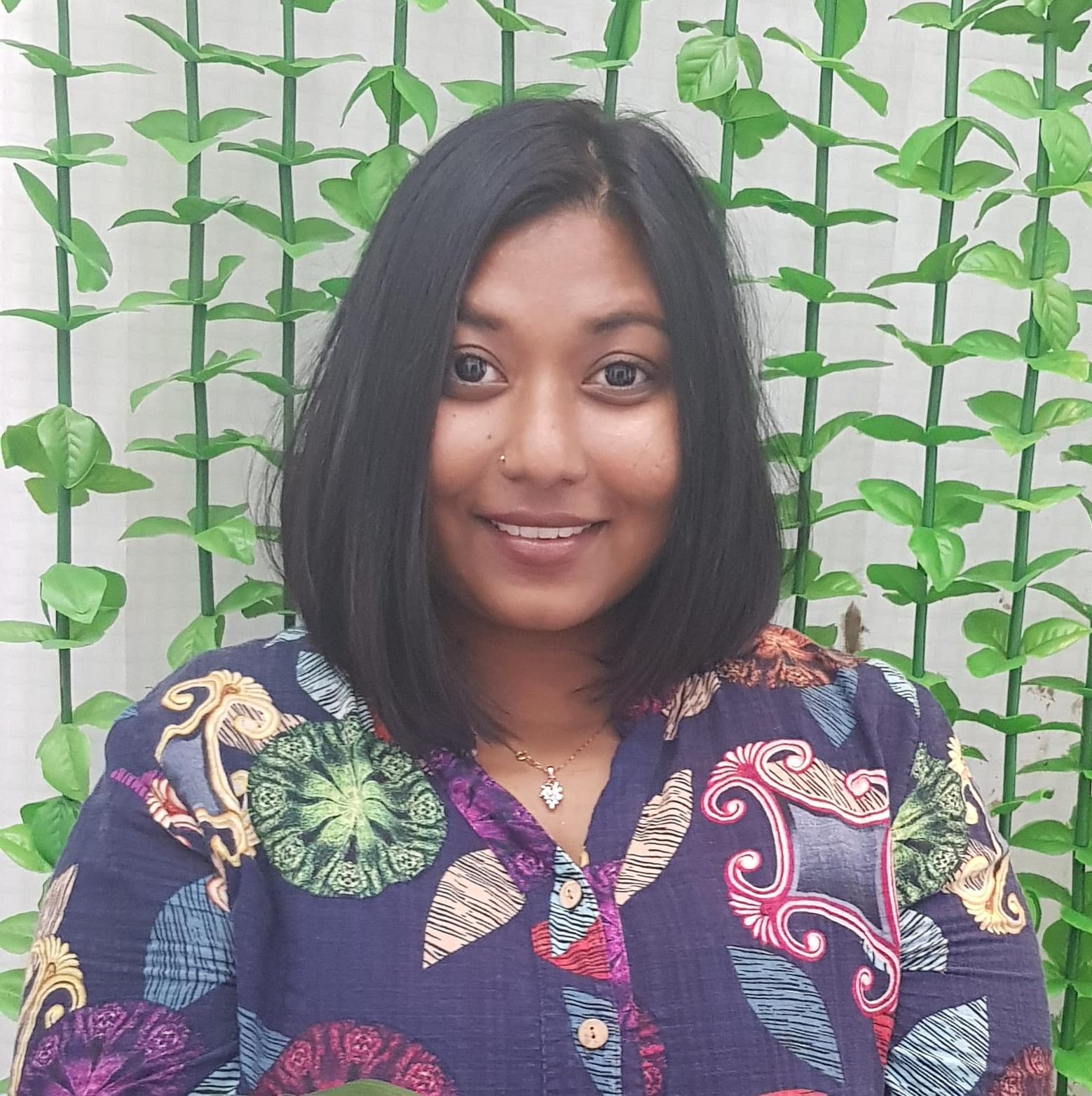 An image of Pramila dressed in a purple patterned shirt standing against a backdrop of leaf vines.