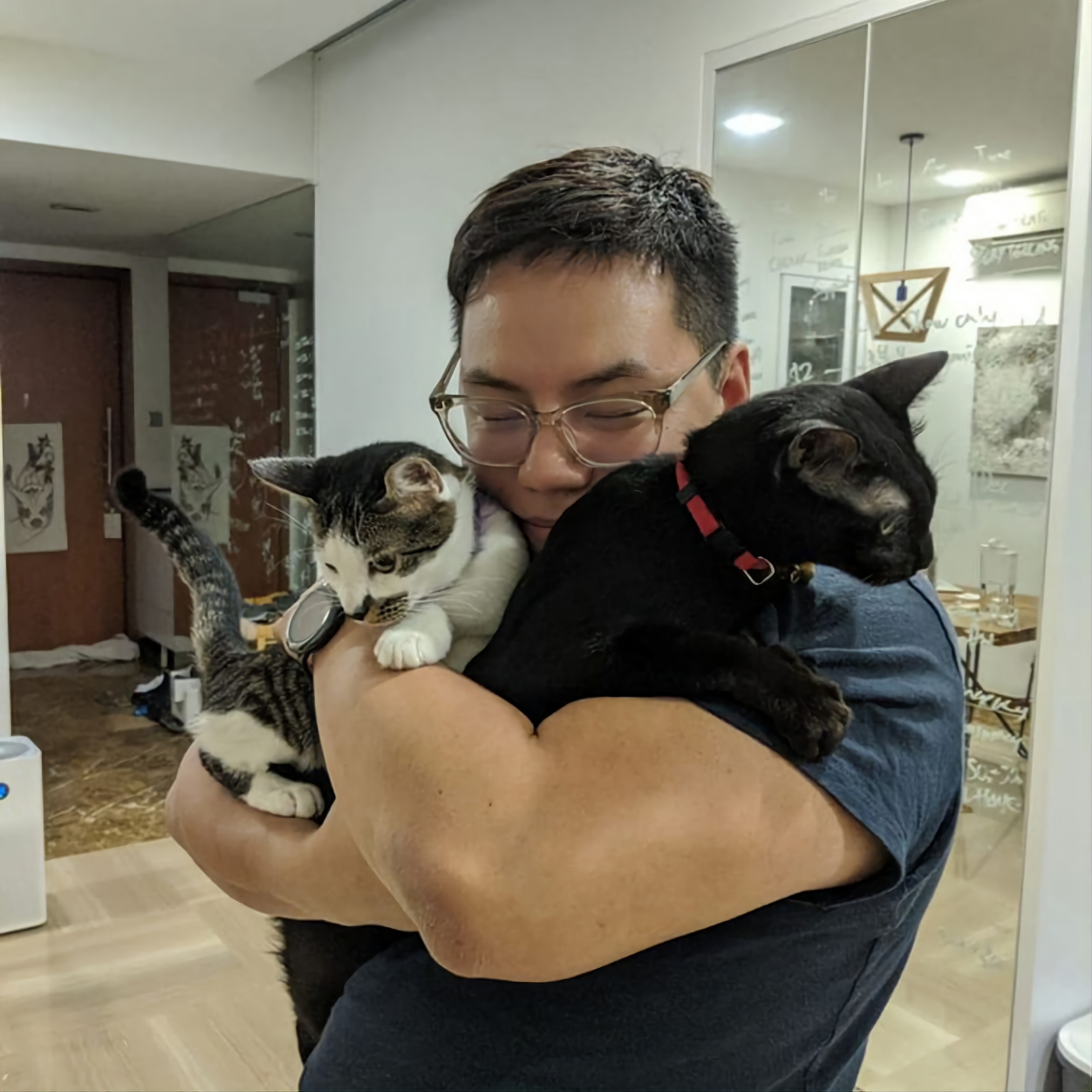 An image of Henrik Cheng dressed in a navy blue shirt cuddling two cats.