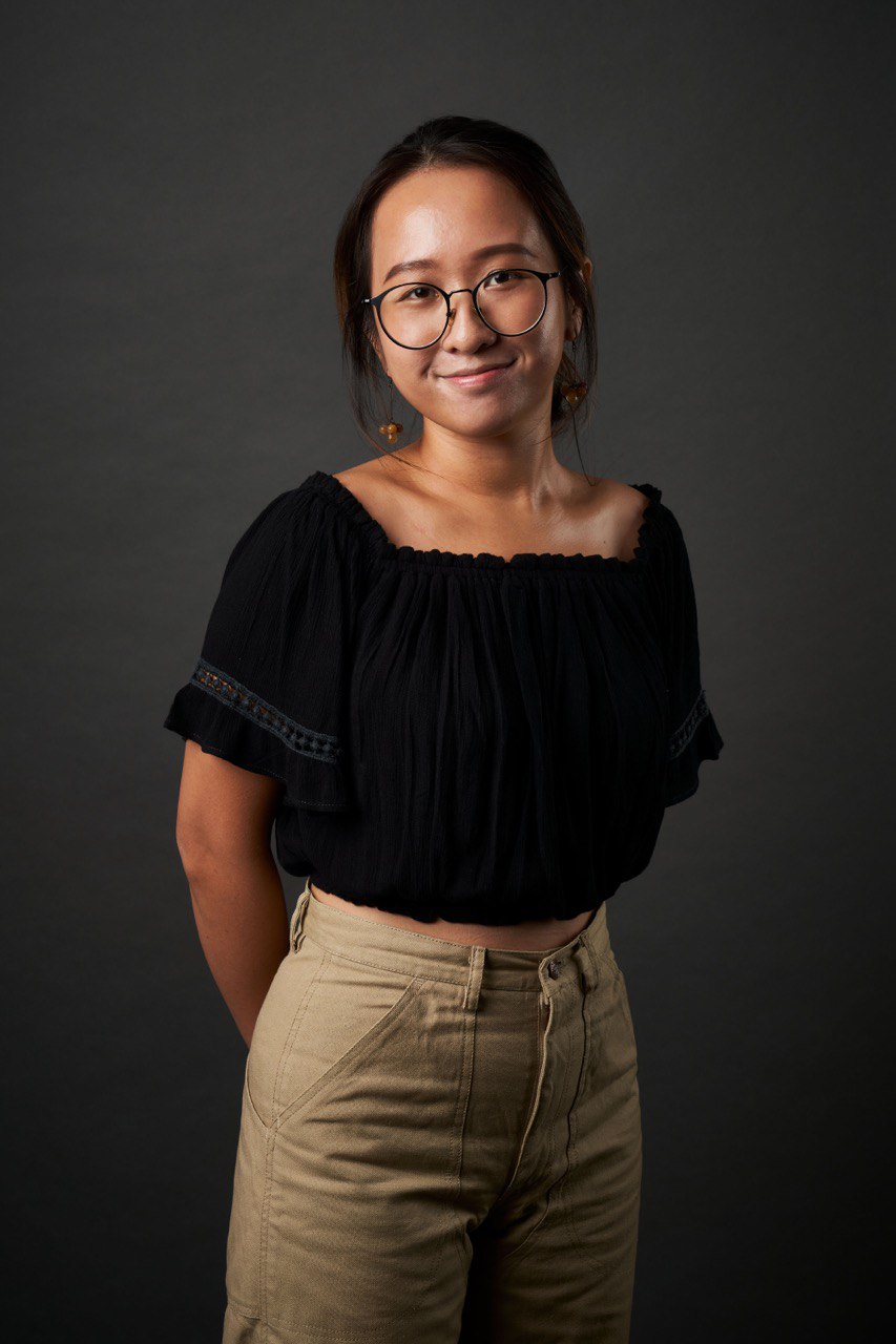 A female figure in a black top with beige pants standing in front of a black backdrop with her hands behind her back