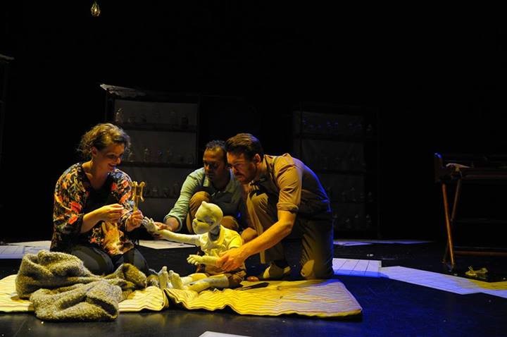 Three persons on a stage, seated and clustered around a small white statue of a boy.