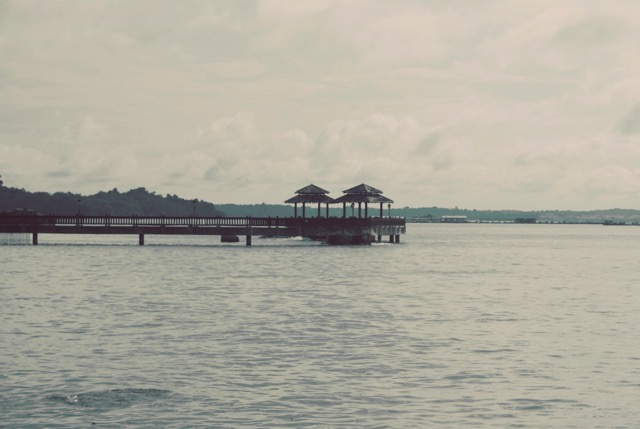 An image of the sea and in the distance a wooden bridge leading up to two sheltered pavilions can be seen.