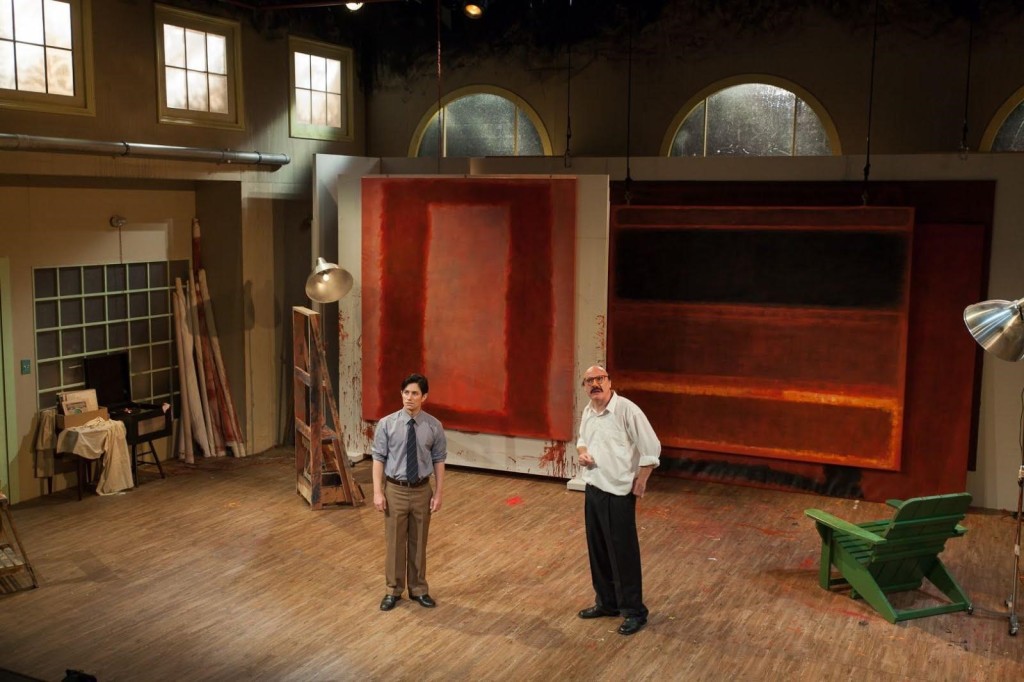 Two male-presenting persons standing on a wooden stage decorated to look like a house.