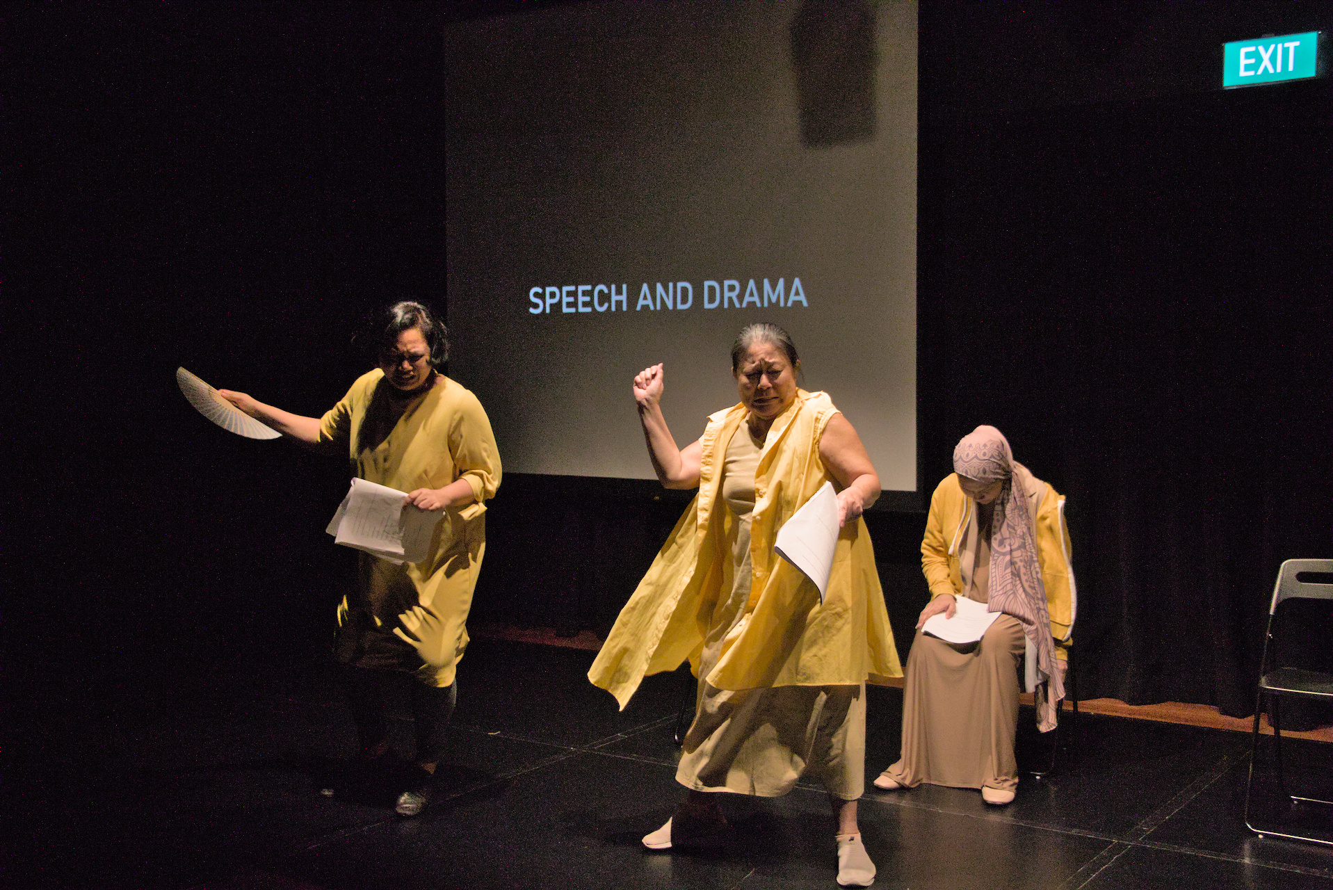 The photograph features three women dressed in yellow clothing, two are standing and are poised in the same act of striking something. The third woman is seated on a chair curled up as if in pain. A screen behind them projects the words "Speech and Drama".