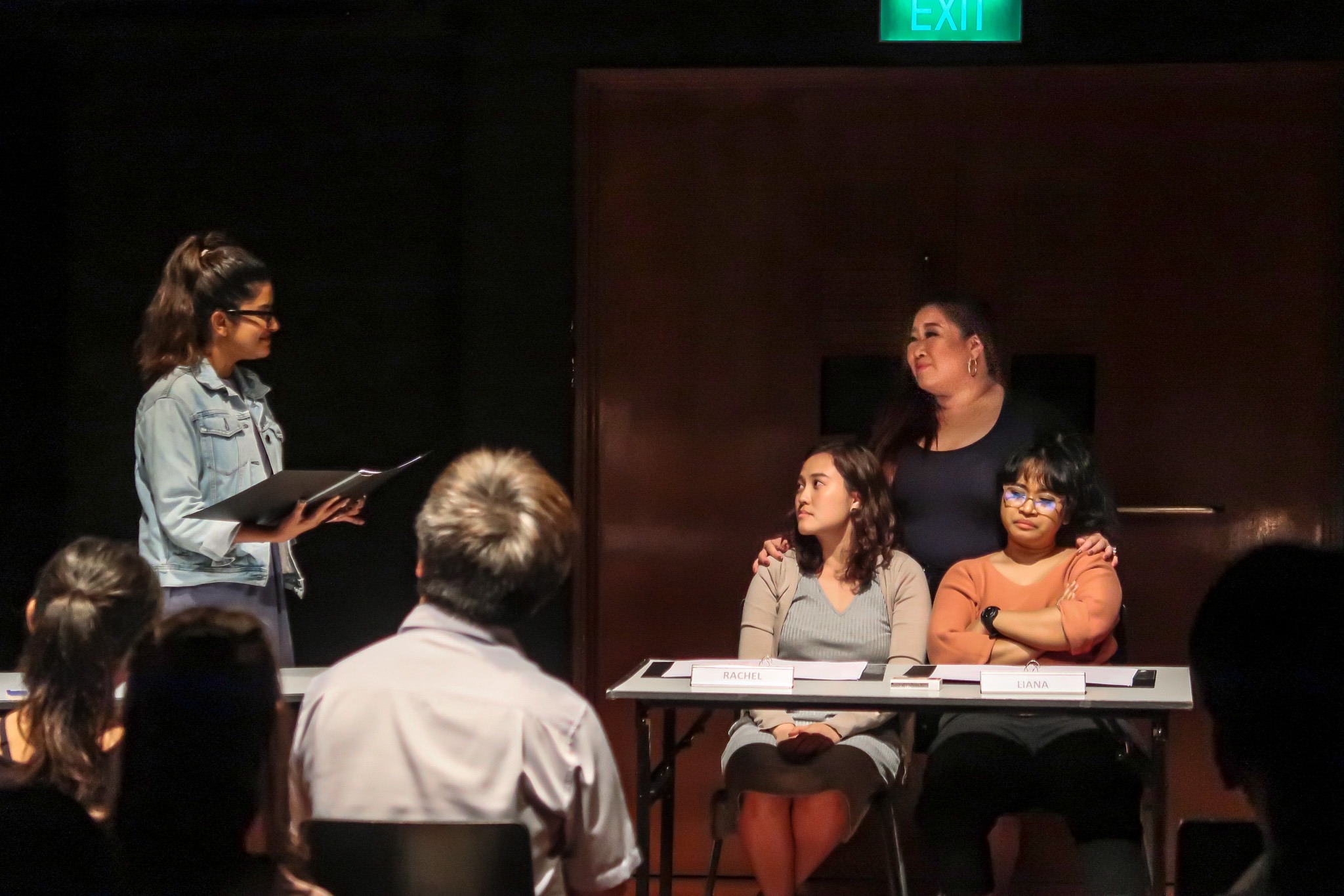 An image in the Black Box with two performers seated in front of a table with scripts on the right and a performer standing behind them looking towards another performer on the left with a black file in hand.