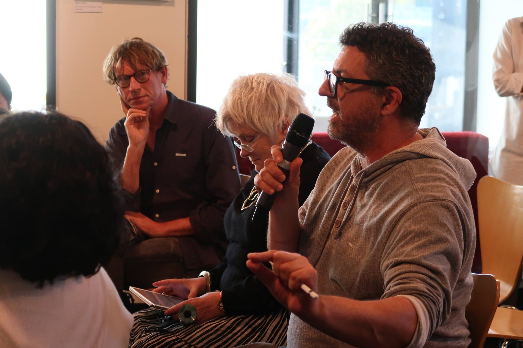 A male-presenting person seated beside two other persons, speaking into a microphone and gesturing with his right hand.