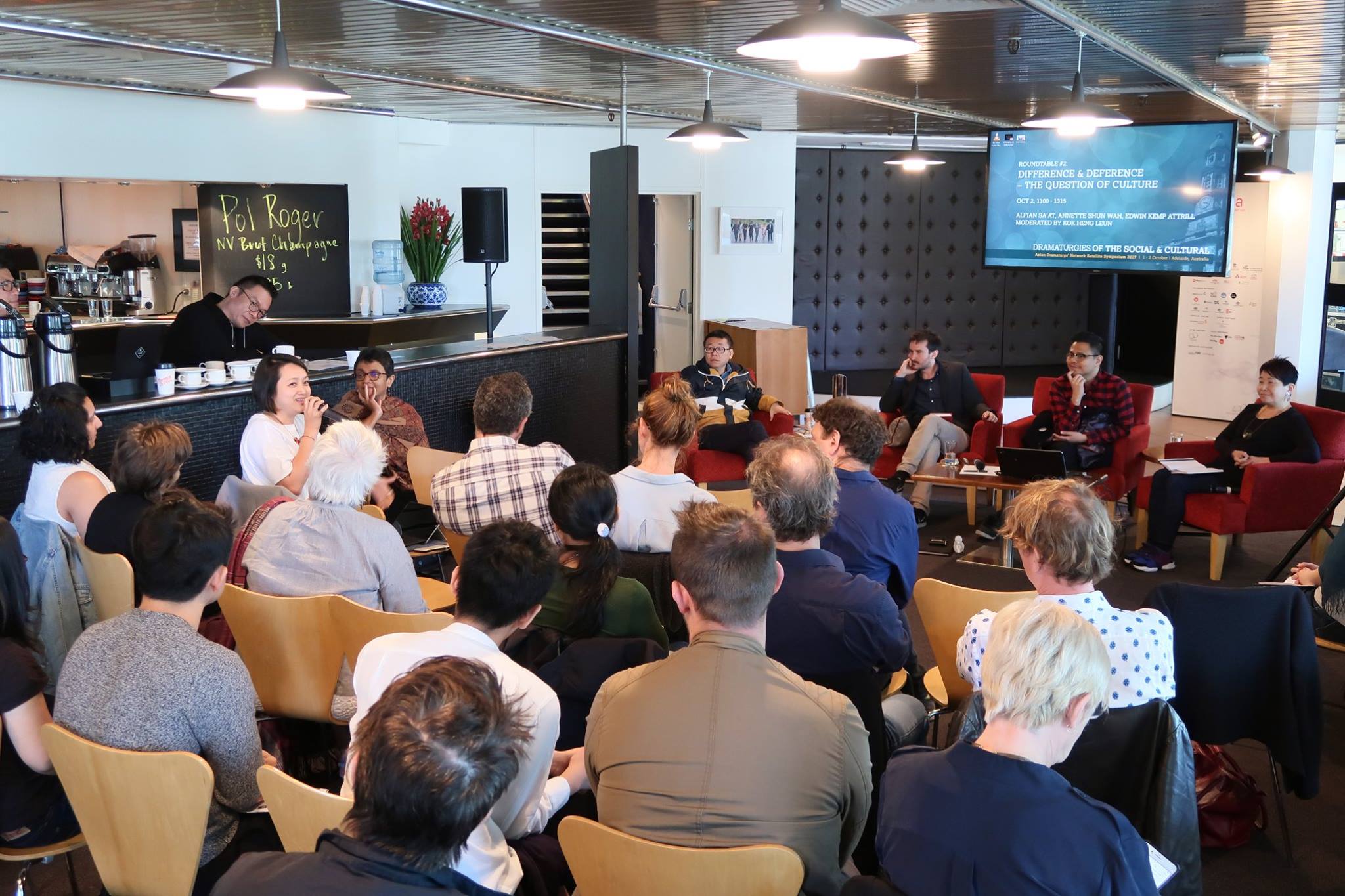 A large audience in a room, looking at a seated person speaking into a microphone.