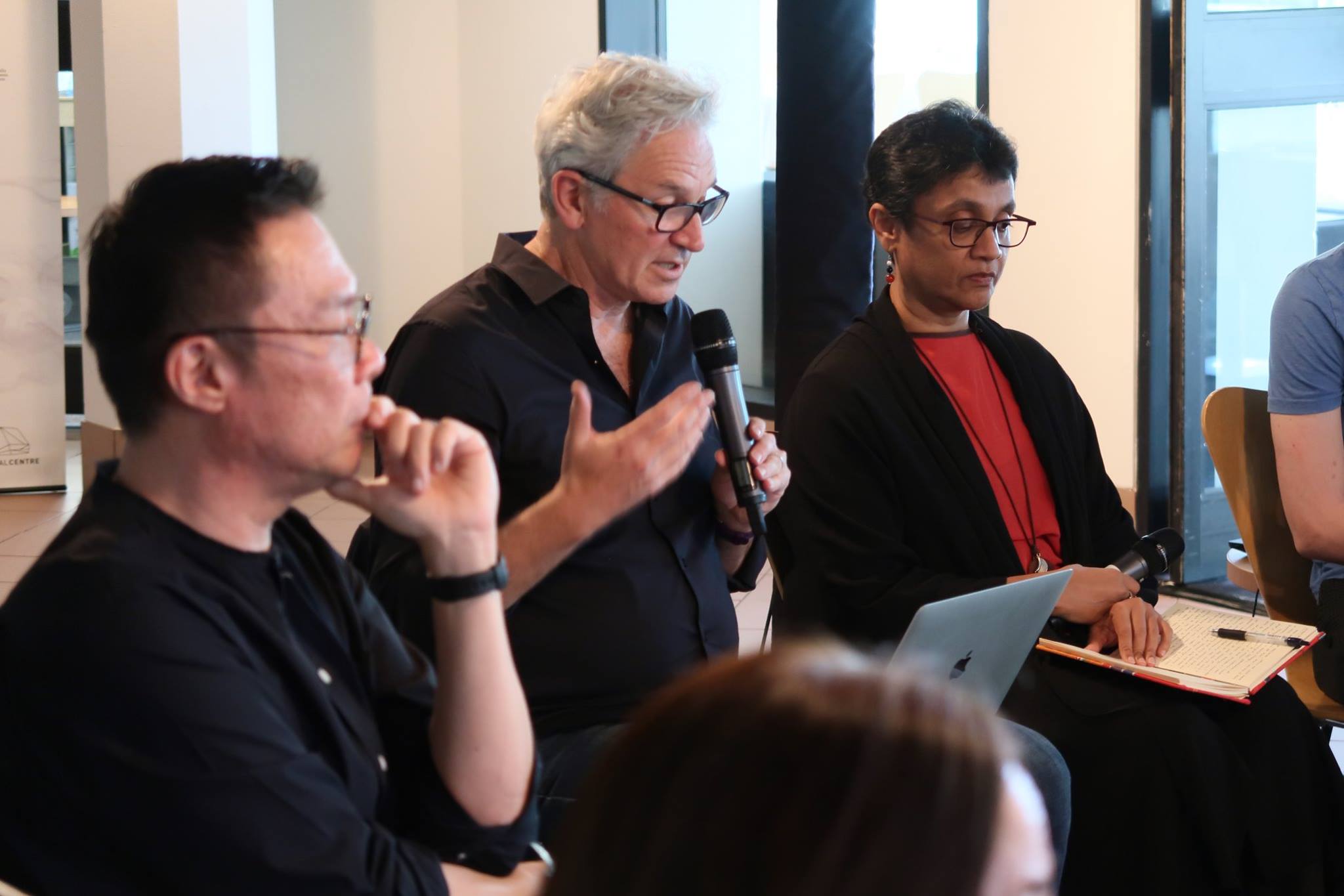 Three persons seated in a row. The person in the middle speaks into a microphone.