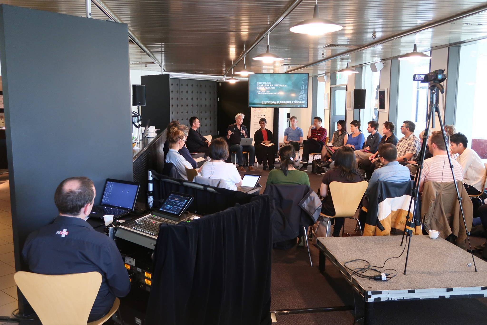 A large group of persons seated in a circle. A video camera is set up on a tripod facing the circle.