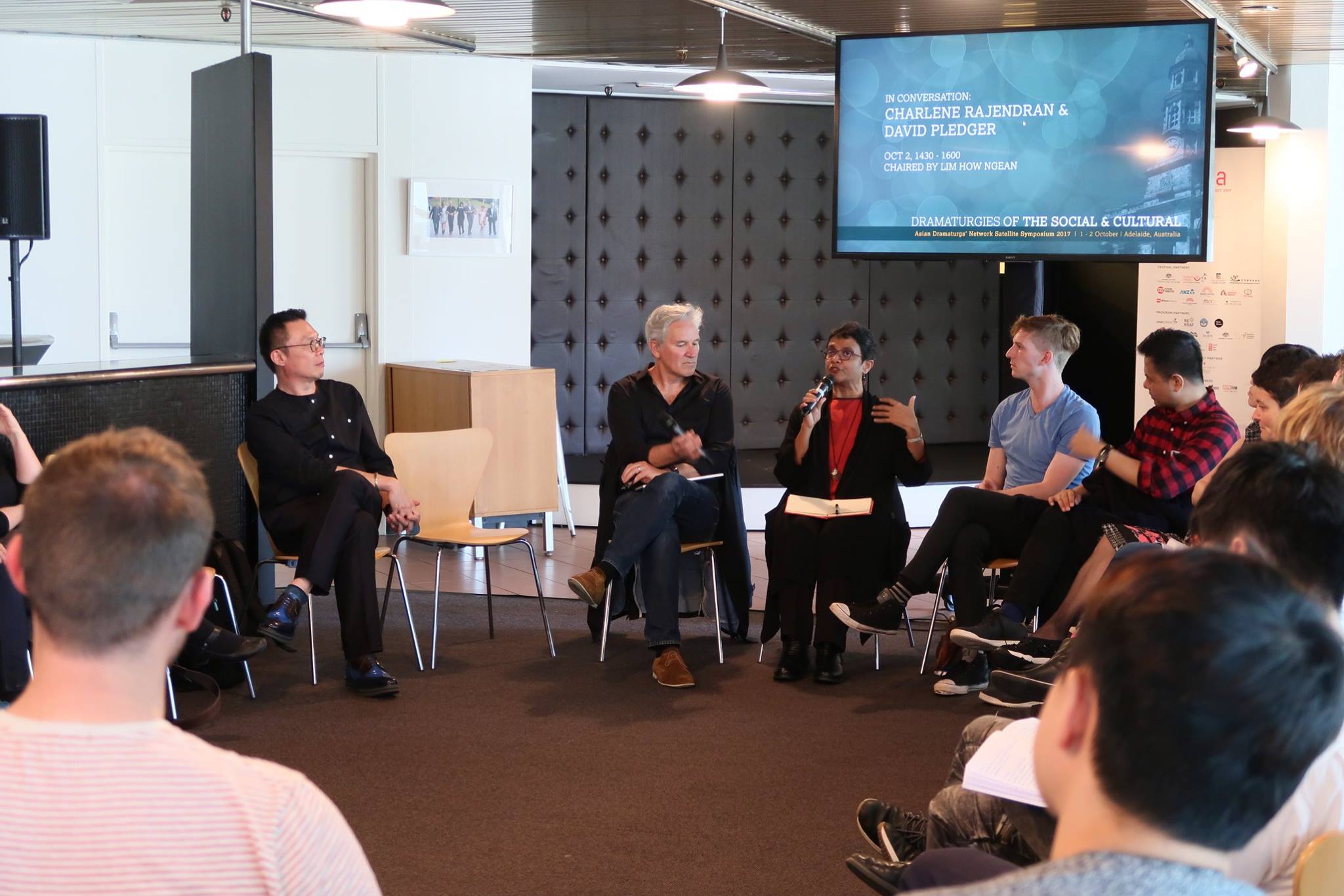 A group of persons seated in a circle, looking at a female-presenting person speaking into a microphone.