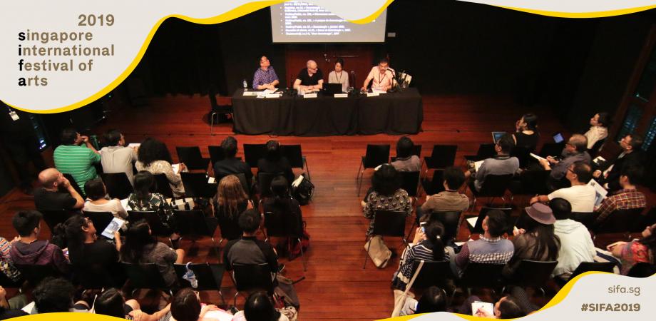 A large audience seated in front of a black table, behind which are four speakers.