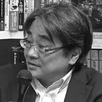 Black and white headshot of Tadashi Uchino speaking into a microphone in front of a shelf of books.