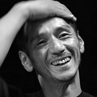 Black and white headshot of Takao Kawaguchi with a hand atop his head, smiling.