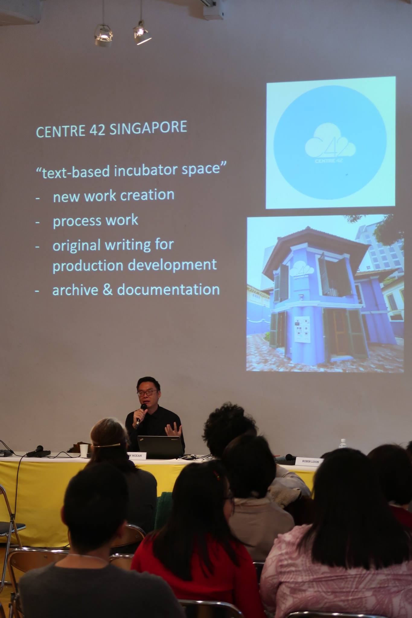 A projector screen with a slide describing Centre 42. A male-presenting person sits in front of the screen and speaks into a microphone.