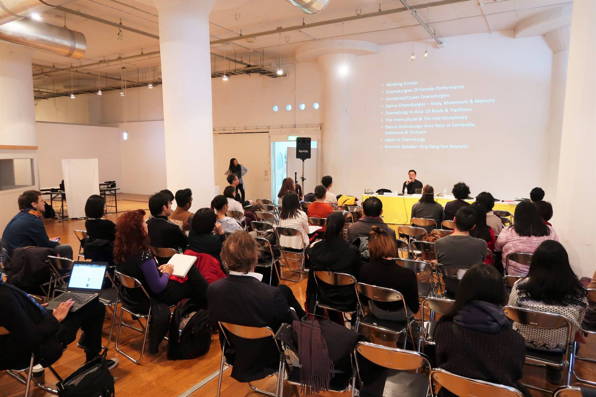 A large audience facing a projector screen with a slide full of text.