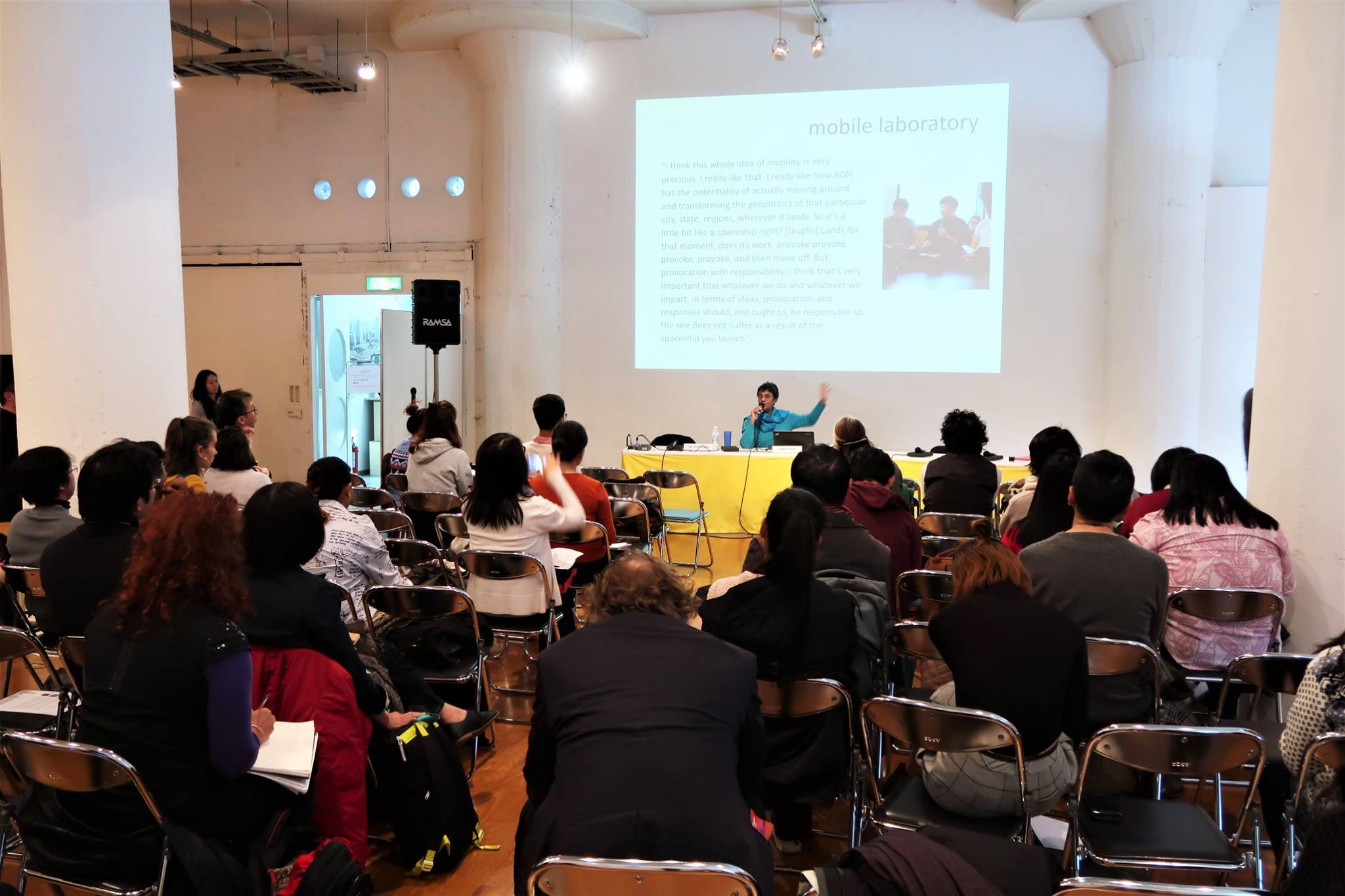 A large audience facing a female-presenting person speaking into a microphone and gesturing at a projector screen.