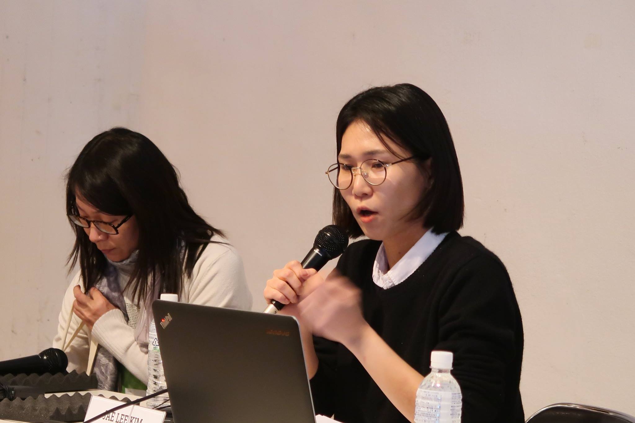 Two female-presenting persons seated side by side. One speaks into a microphone while looking at her laptop.