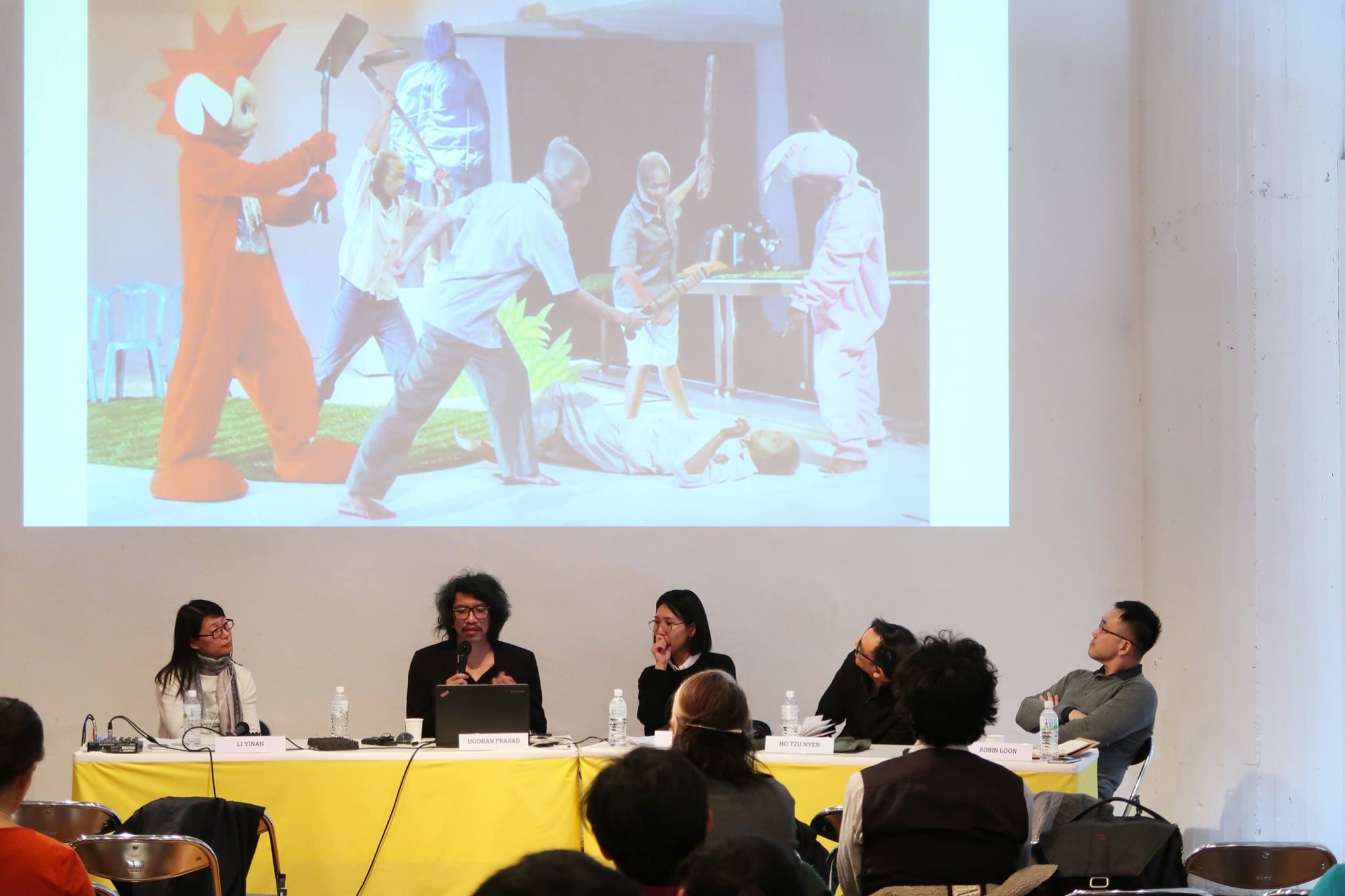 Five persons seated at a long table. Above them is a projector screen depicting a scene of violence.