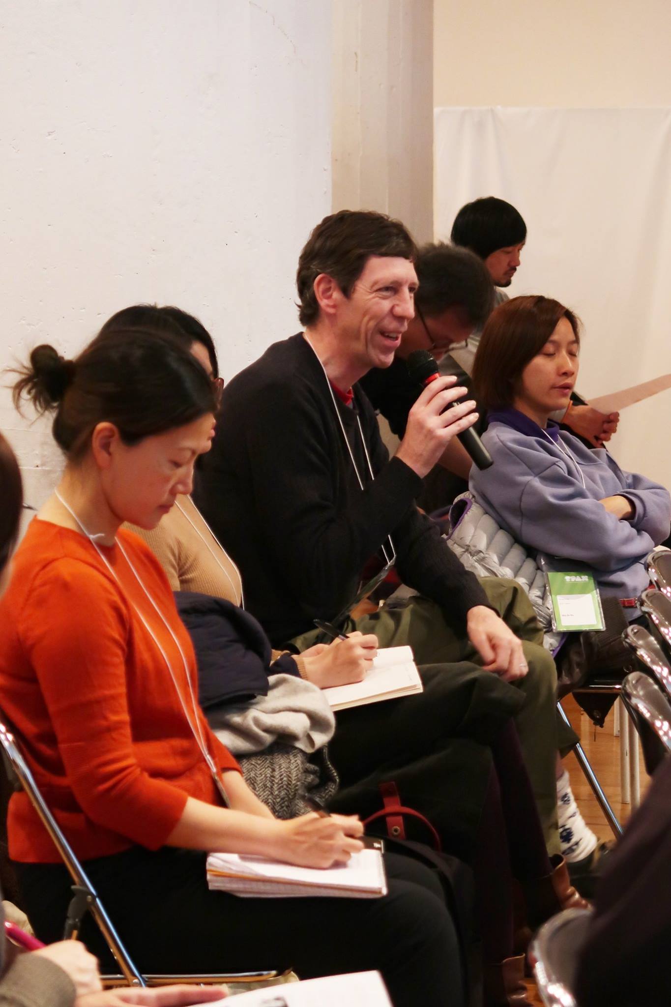 A row of persons seated in the audience. A male-presenting person speaks into a microphone.