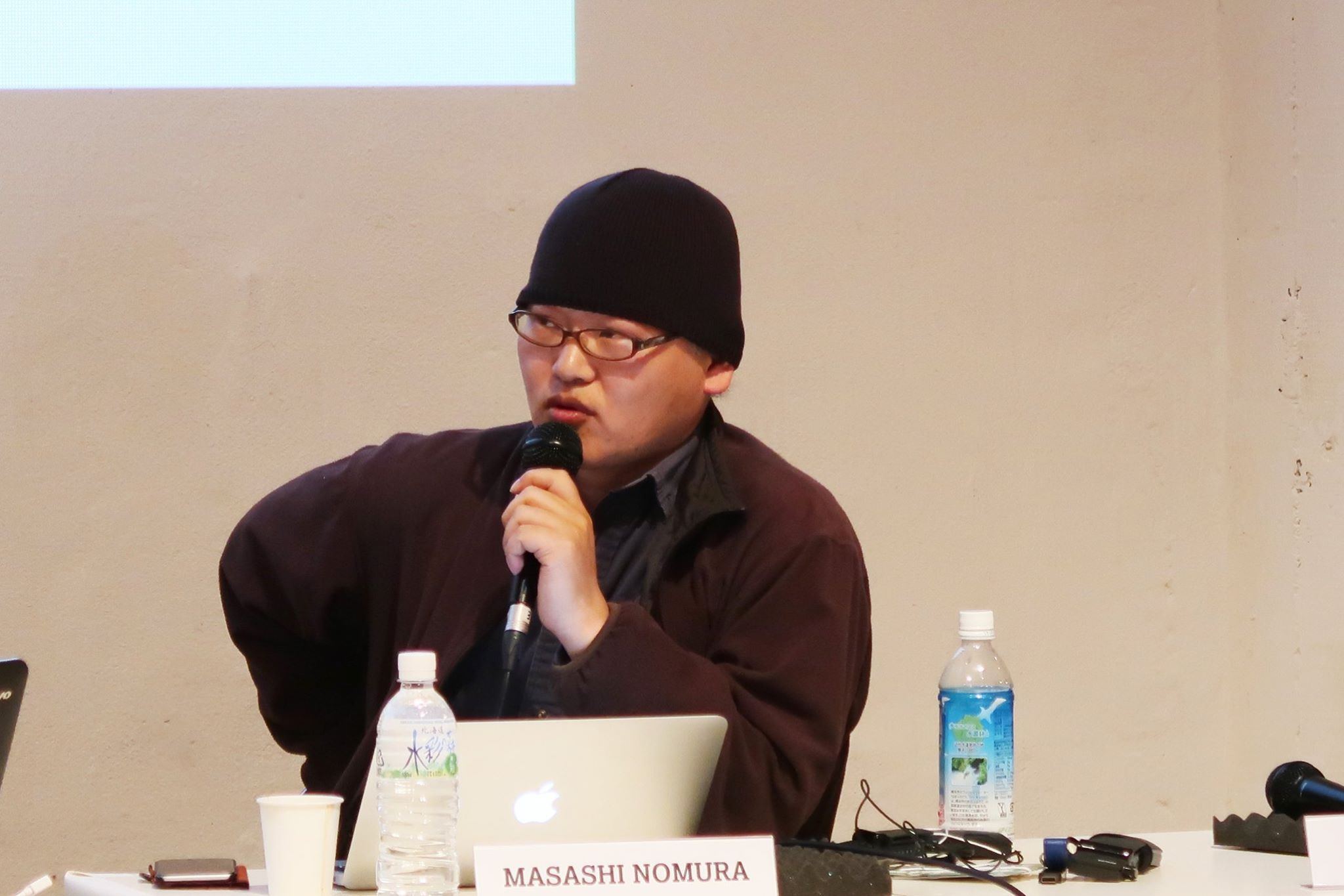 A male-presenting person in glasses and a beanie, speaking into a microphone.