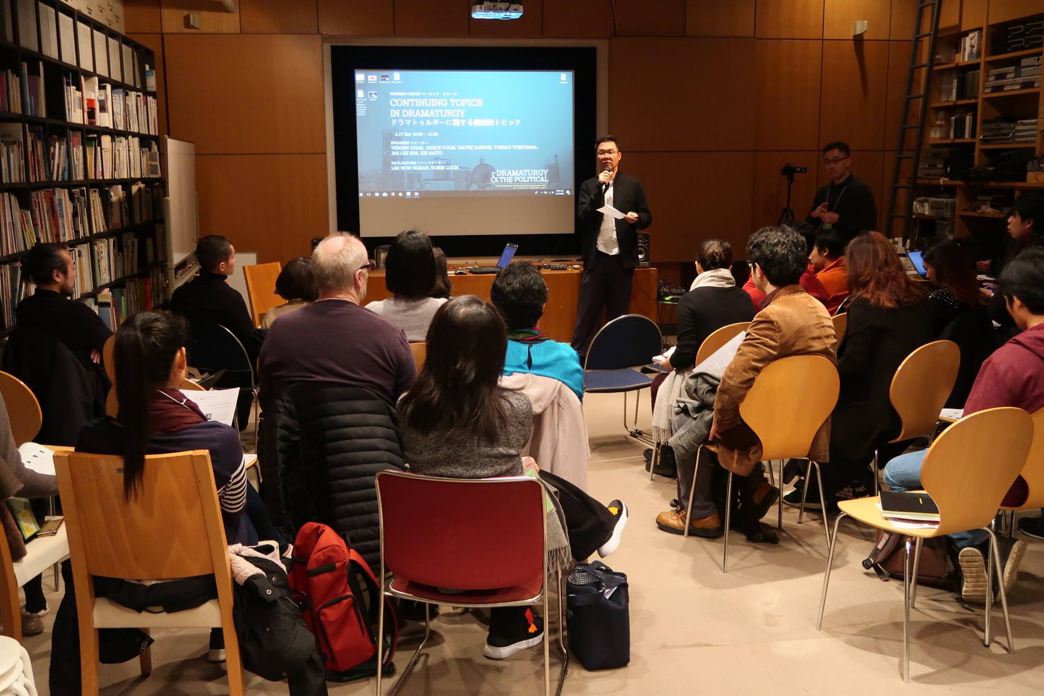 An audience in a library watching a male-presenting person speak into a microphone.