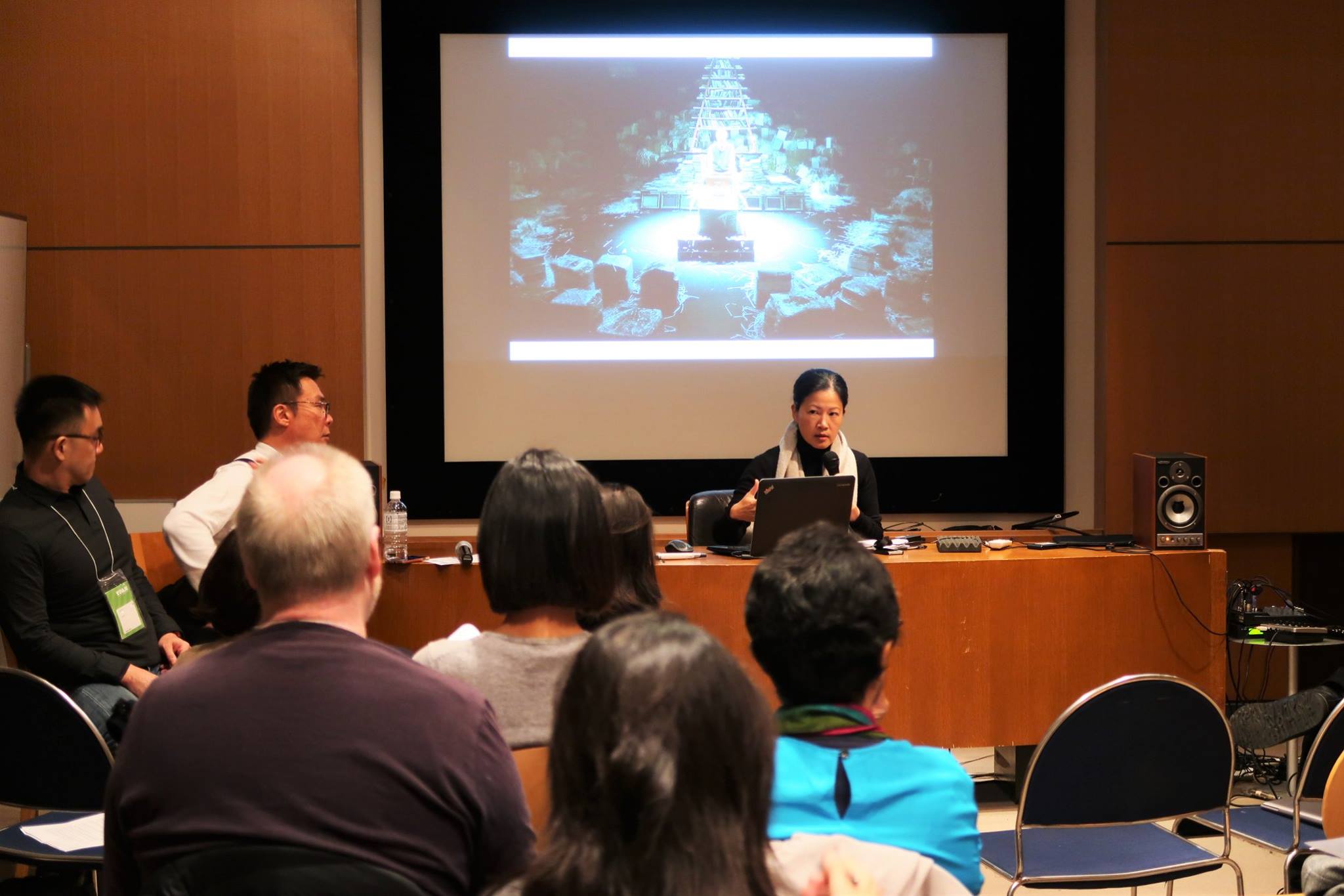 A female-presenting person seated at a long table, facing an audience and speaking into a microphone.