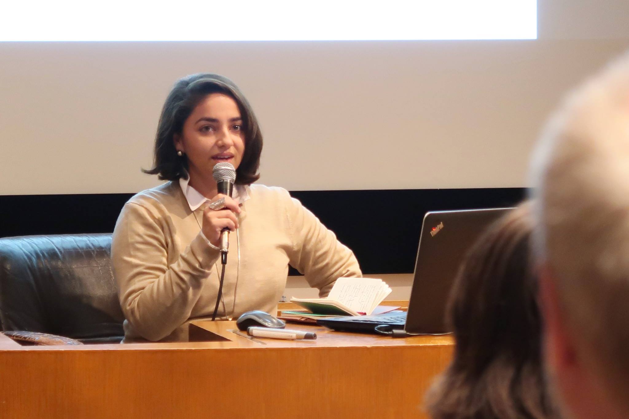 A female-presenting person in a yellow sweater speaking into a microphone.