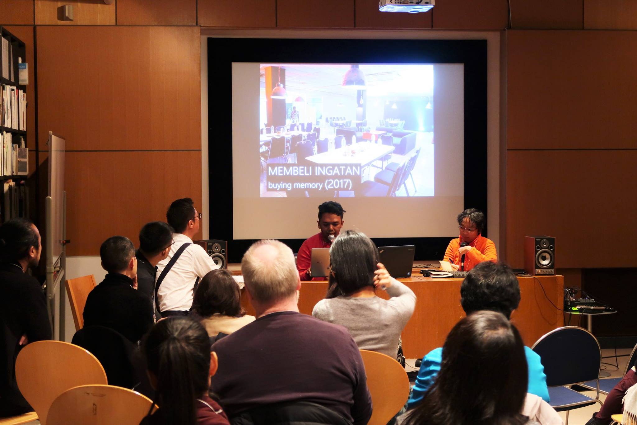 Two persons seated at a long table, speaking into microphones. Behind them is a projector screen depicting the interior of a restaurant.