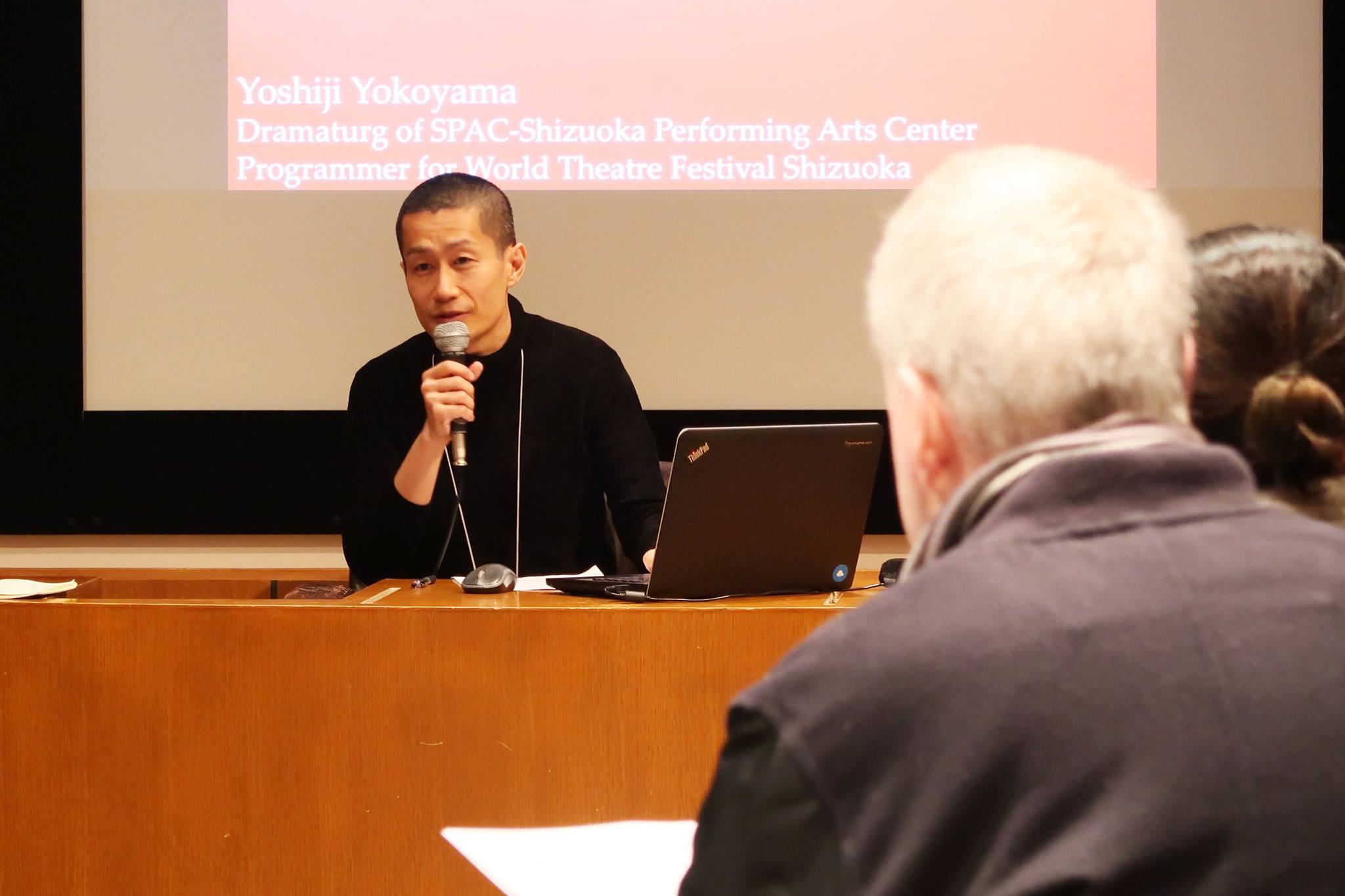 A male-presenting person in a black jacket, speaking into a microphone.