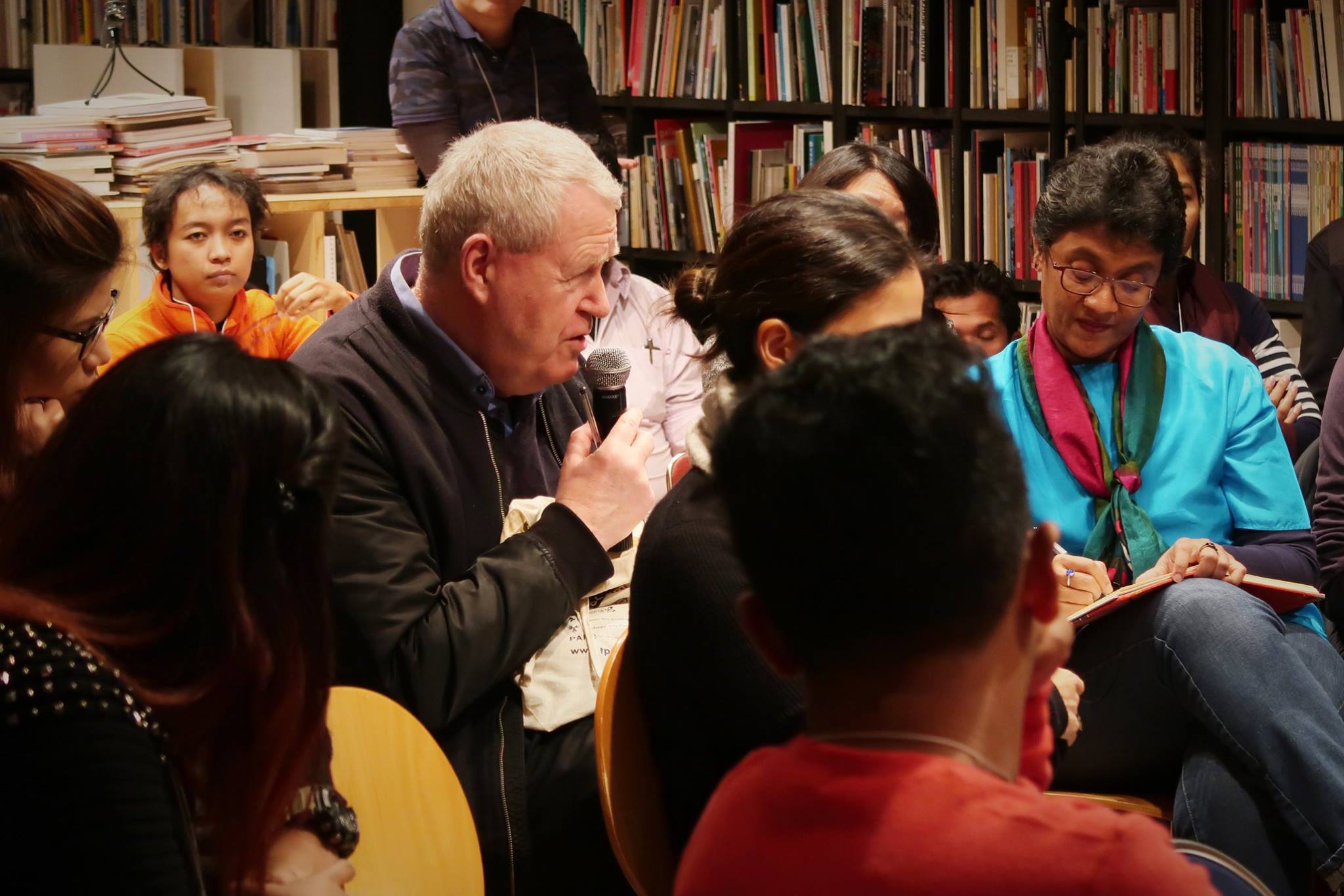 A crowded audience. One male-presenting person speaks into a microphone.