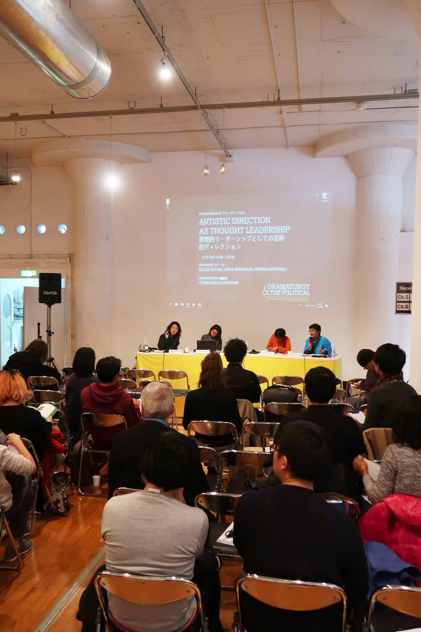 A large audience facing a long table, at which four persons are seated.