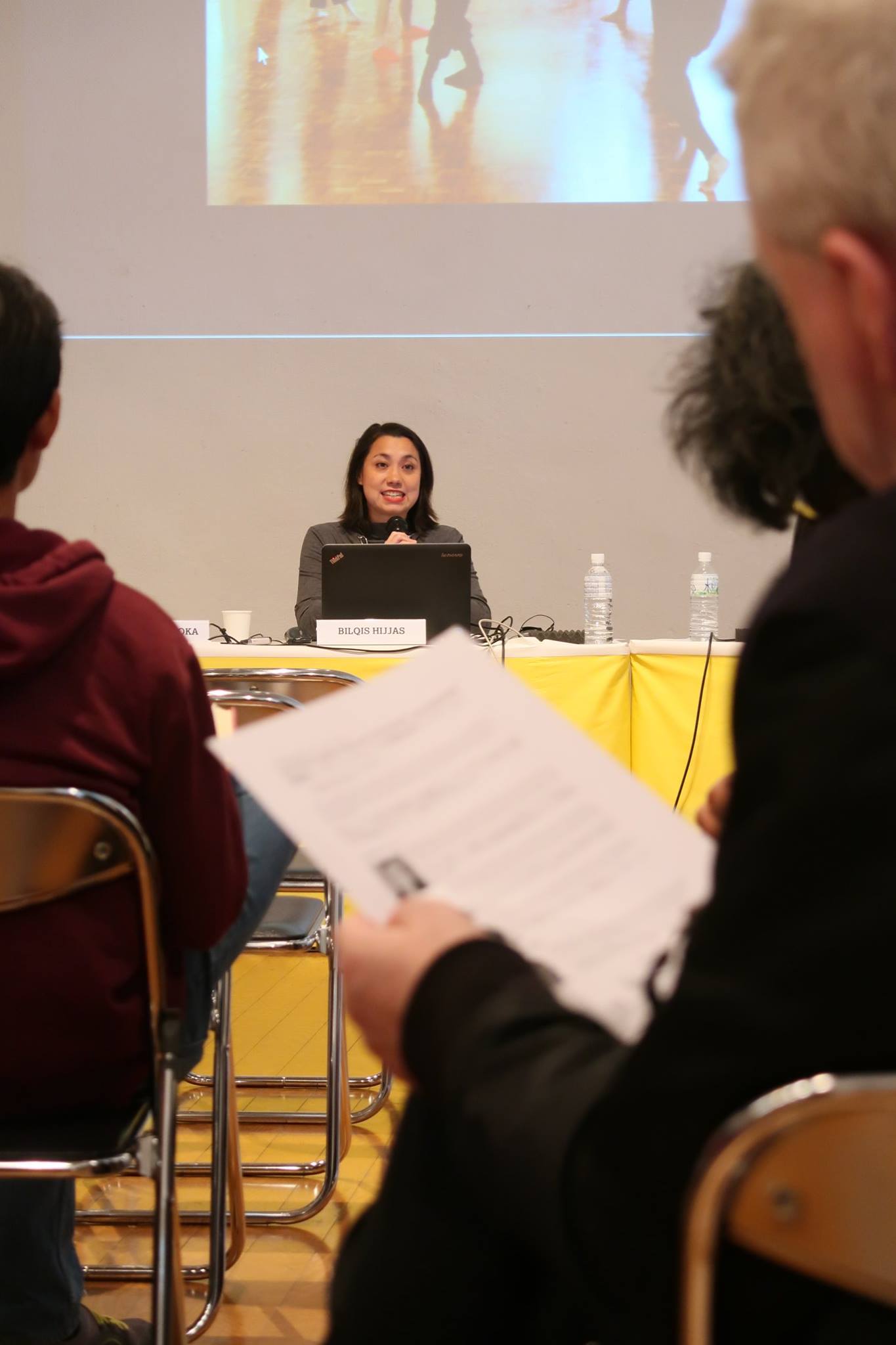 A female-presenting person smiling and speaking into a microphone while facing an audience.