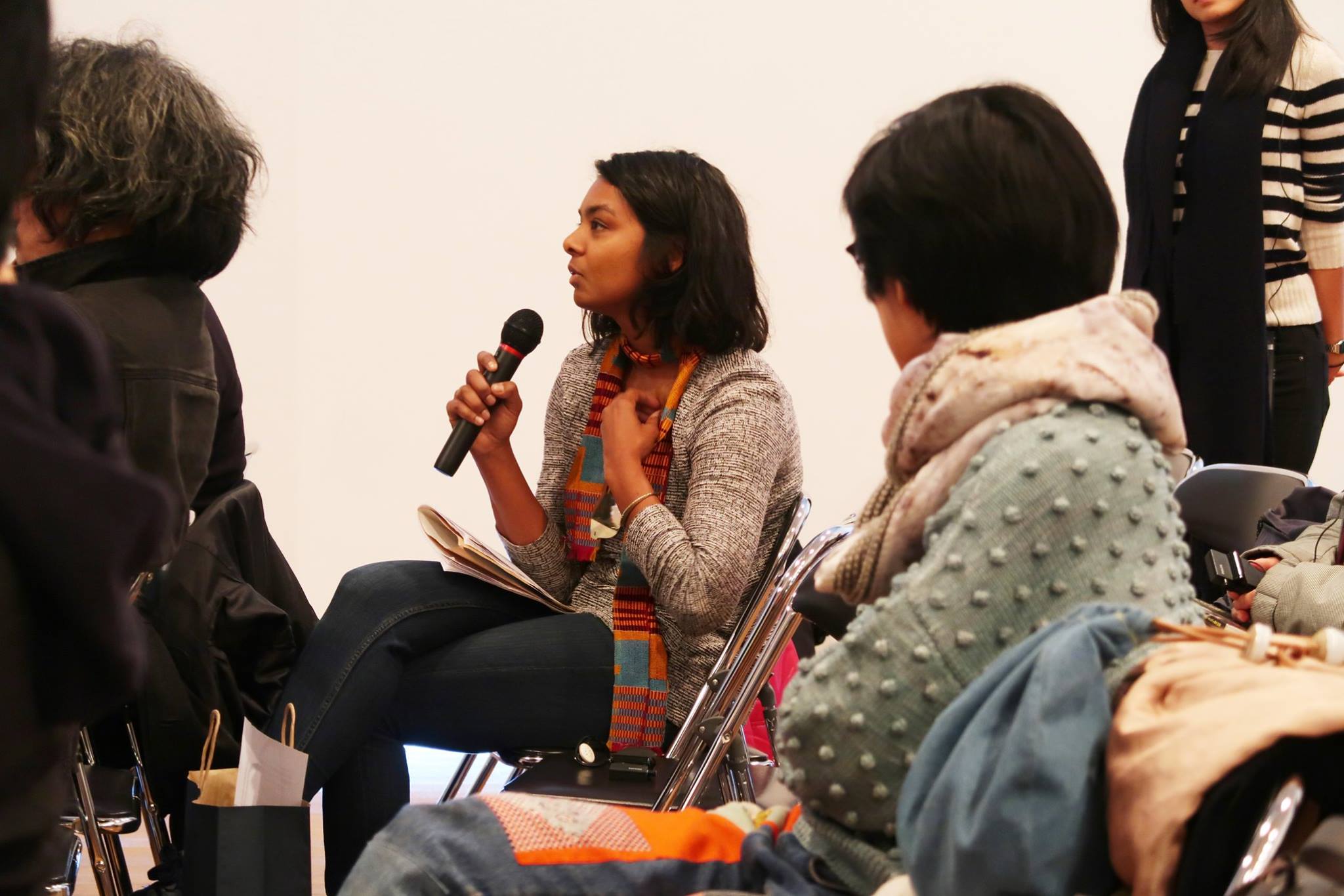 A female-presenting person speaking into a microphone amidst an audience.