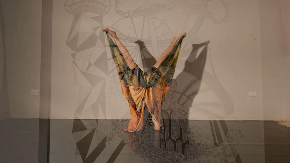 A person draped in a colourful cloth doing a handstand, legs spread wide. Overlayed on them is a translucent geometric image.