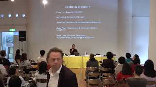 An audience sitting in front of a long yellow table and projector screen.