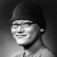 Black and white headshot of Masashi Nomura, smiling, wearing glasses and a beanie.