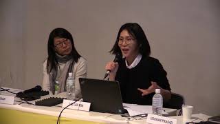 Two female-presenting persons seated at a yellow table. One speaks into a microphone.