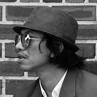 Black and white profile headshot of Ugoran Prasad wearing a hat and mirrored glasses.