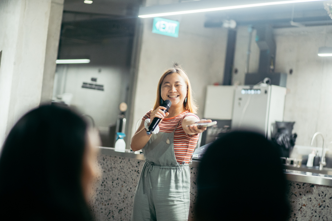 Cheryl Ho in denim overalls, holding a microphone, smiling with right hand outstretched.