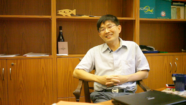 Tan Tarn How in a short-sleeved shirt, sitting in front of some wooden shelves, hands clasped in front of him and smiling.