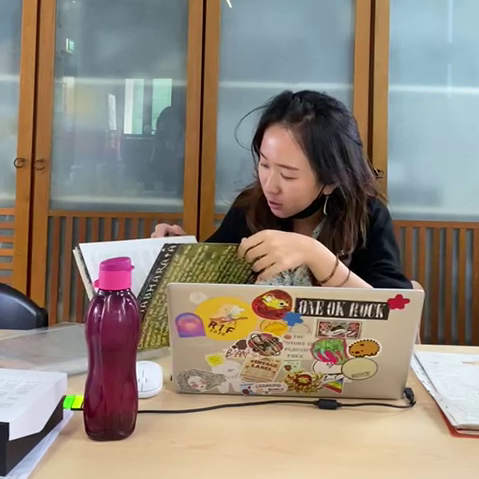 A female-presenting person seated in front of a laptop, looking through a programme booklet.