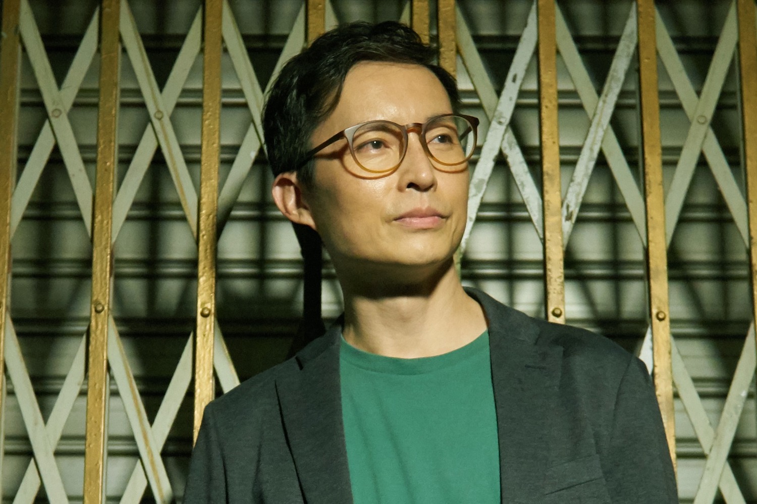 Half-body shot of Nelson Chia in front of a metal grate, wearing a green shirt and glasses, glancing to the right.